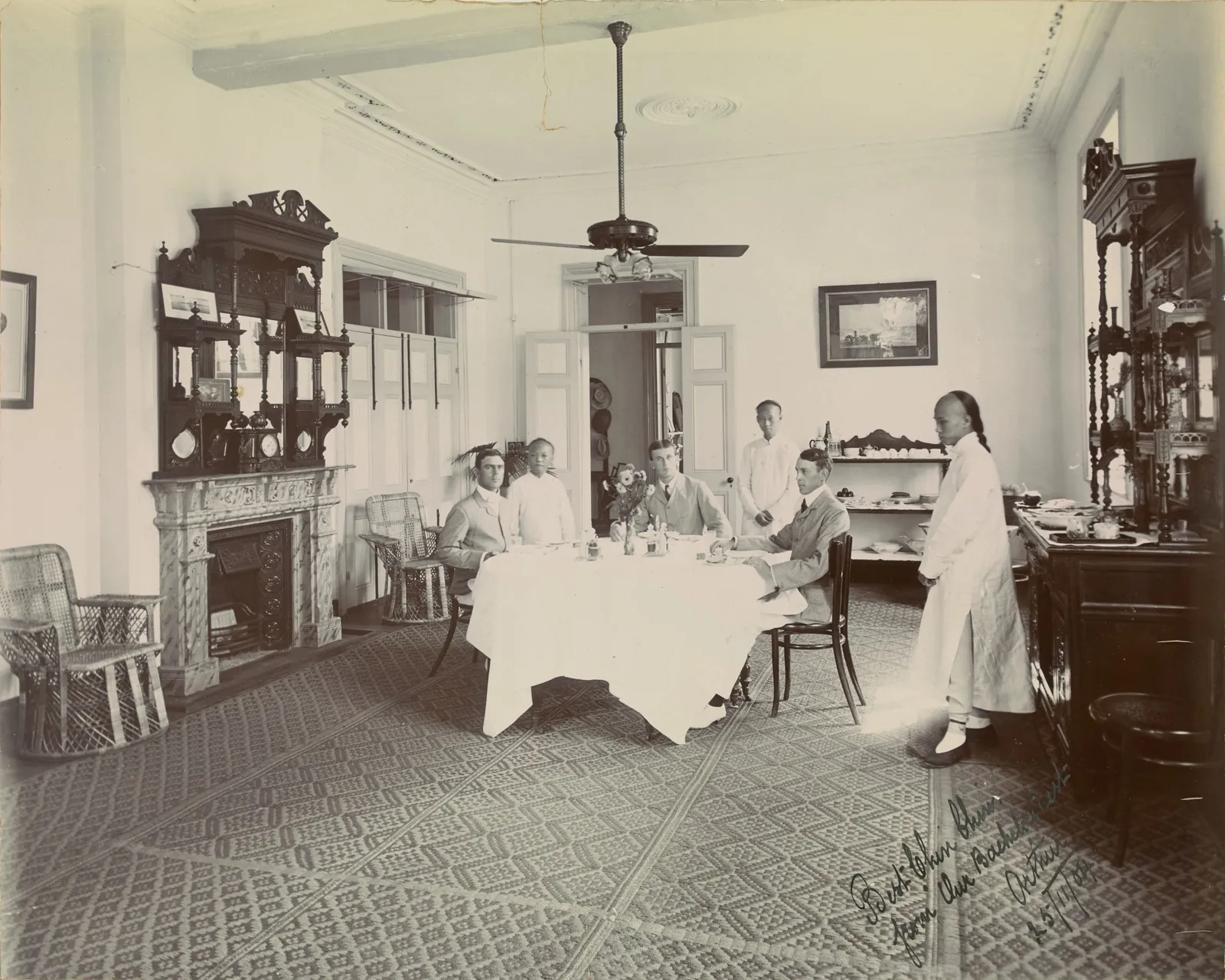 A high dining room with a ceiling fan, a marble fireplace and carved cabinets. Three men sit at a round table and three Chinese servants stand.