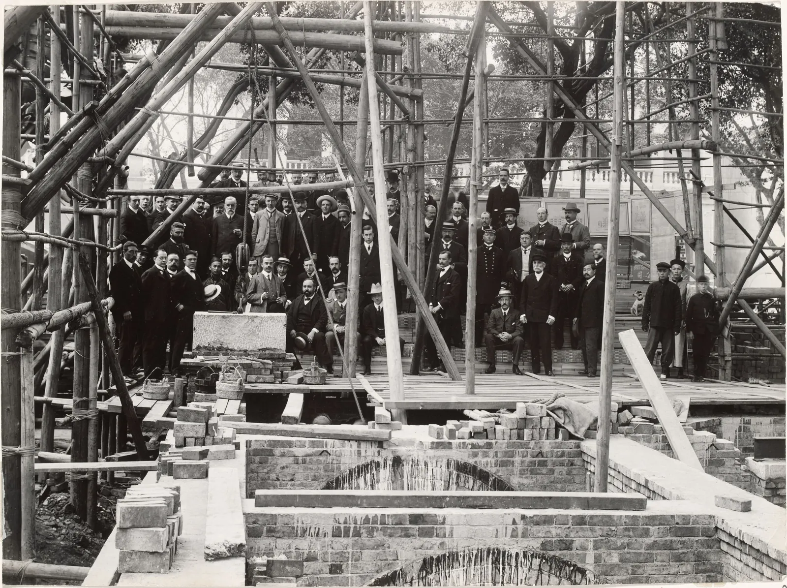 A crowd of men in suits among bamboo scaffolding, above brick foundations and an arch.