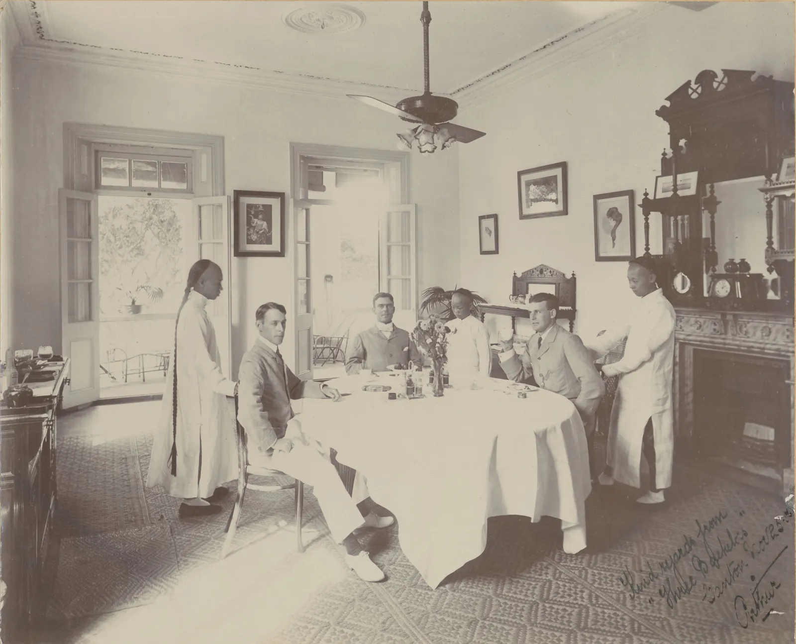 Three men at a round breakfast table under a ceiling fan, with two servants in long white gowns standing by and French doors open to a verandah.